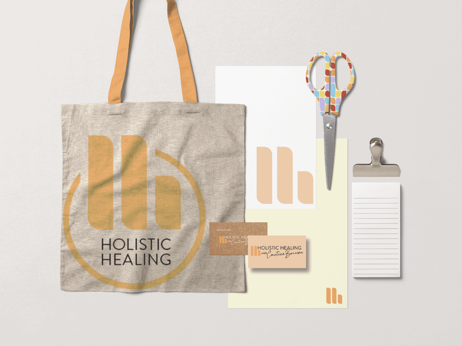 A collection of branded items for Holistic Healing is shown, including a beige tote bag, a pair of scissors with a colorful handle, a business card, a letterhead, a notepad, and a clipboard. The items feature a logo with the letter "H" in an orange and beige color scheme. - Market Design Team: Define. Structure. Expand.