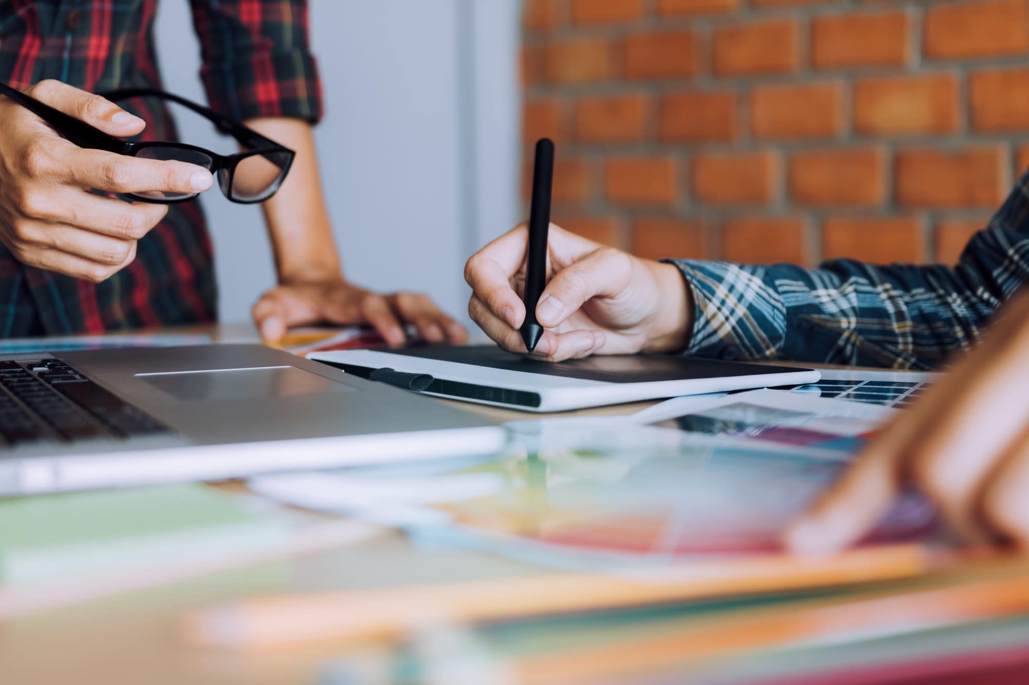 Two people working at a desk. One person holds glasses while the other uses a stylus on a tablet, collaborating on creative solutions. There are a laptop and various documents and color samples on the desk. - Market Design Team: Define. Structure. Expand.