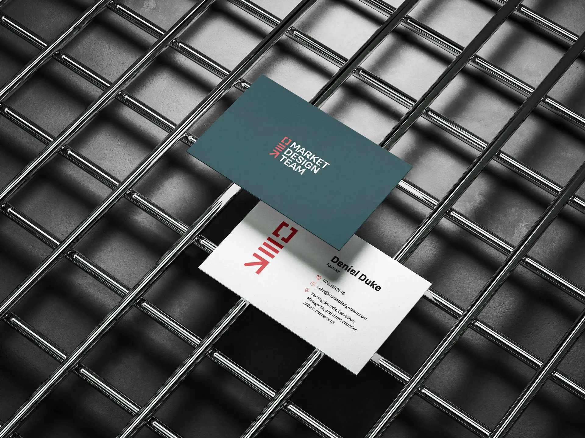 Two business cards resting on a metallic grid surface. One card is dark green with white and red text, displaying "Market Design Team". The other card is white with black and red text, showing contact information for "Daniel Duke" along with a logo, displayed in the "About Us" section.