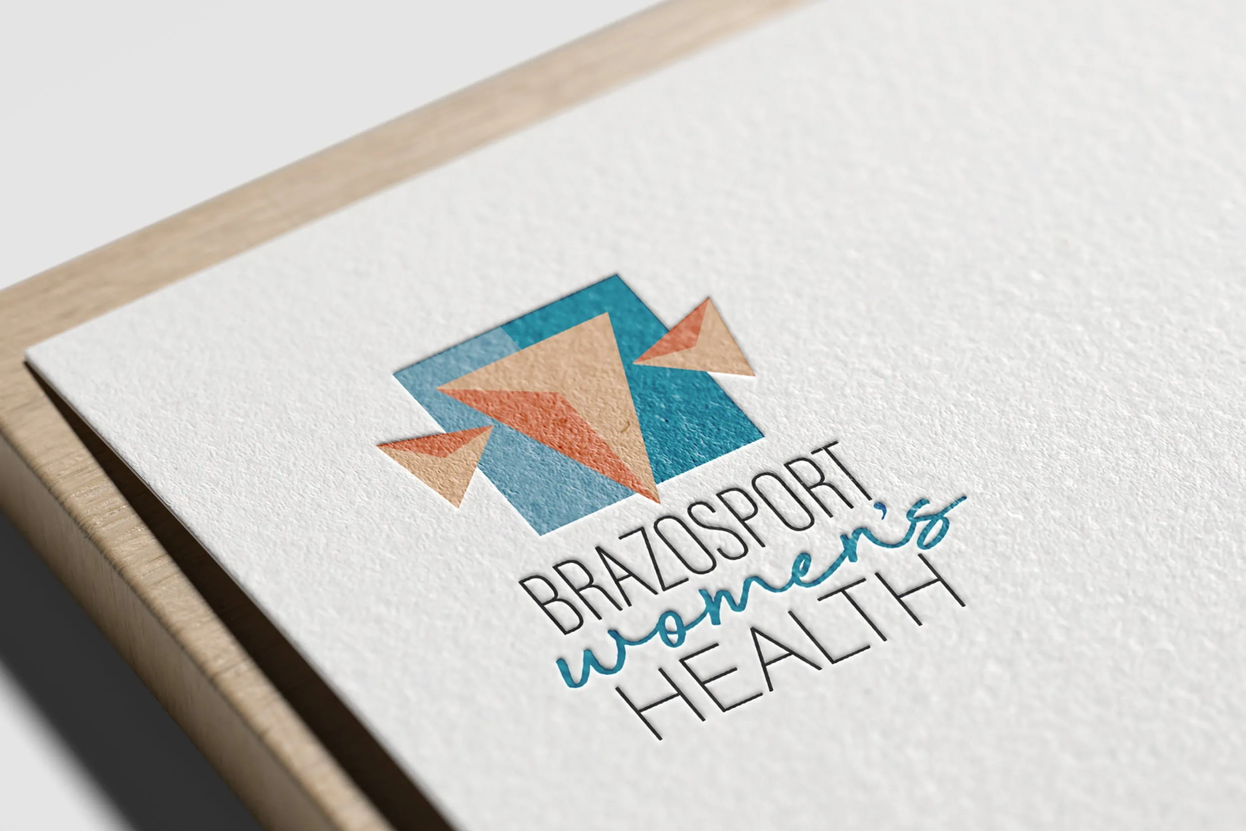 A close-up image reveals a textured white paper featuring the Brazosport Women's Health logo, composed of geometric shapes in blue and orange. The paper rests elegantly on a light wood surface. - Market Design Team: Define. Structure. Expand.