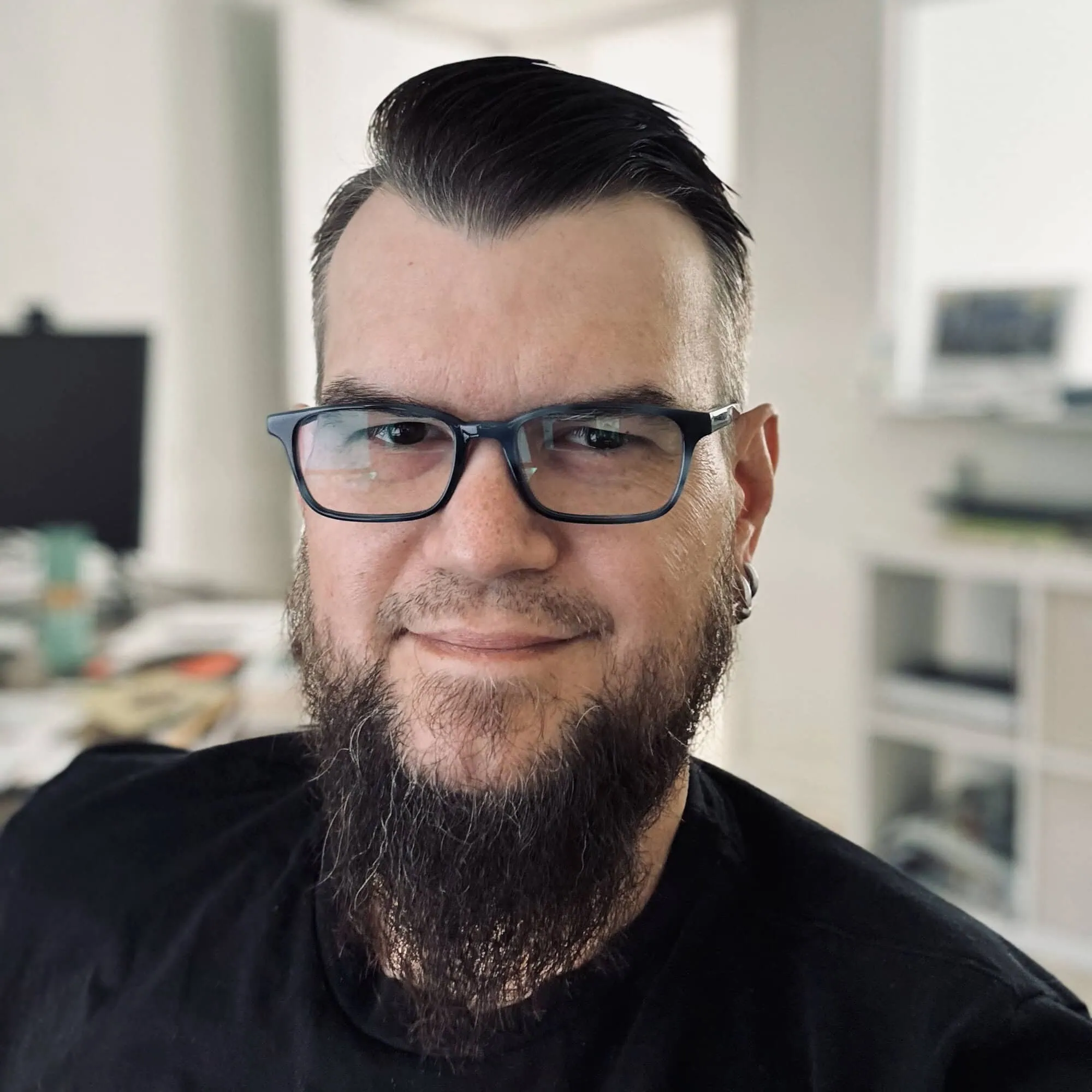 Daniel Duke, a man with short, dark hair and a long, full beard, is wearing black-framed glasses and a black shirt. He is indoors in a room with shelves and a computer visible in the background. He's smiling slightly at the camera. - Market Design Team: Define. Structure. Expand.