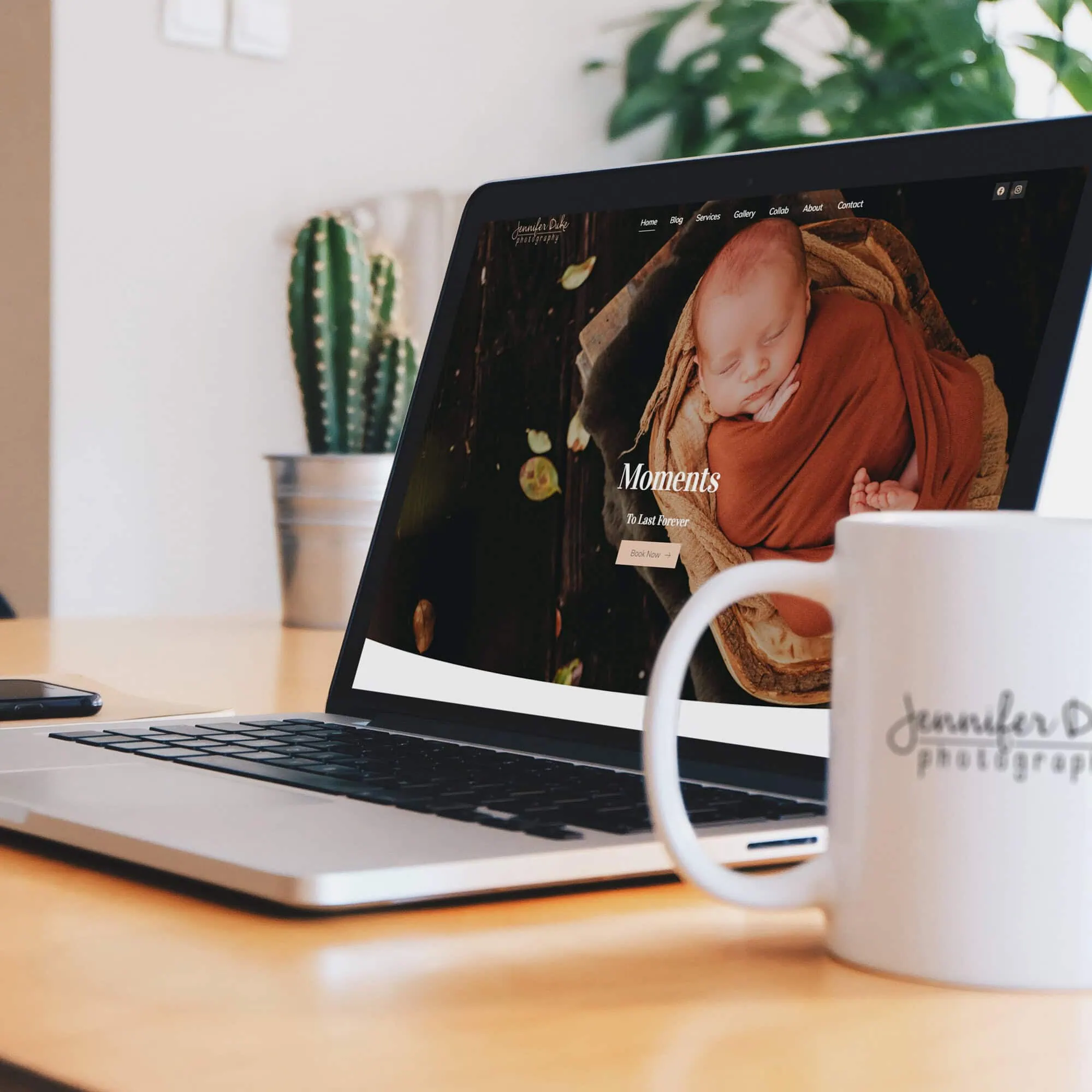 A laptop displaying a website with a photograph of a sleeping baby wrapped in a red blanket. The laptop is placed on a wooden desk near a white mug with "Jennifer Duke Photography" printed on it. A cactus plant is in the background. - Market Design Team: Define. Structure. Expand.