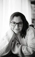 Black and white photo of Jennifer Duke with long, wavy hair, wearing glasses and a knitted sweater. She is resting her chin on her hand, gazing at the camera. The background is blurred, with light-colored curtains nearby, showcasing Duke's expertise in portrait photography. - Market Design Team: Define. Structure. Expand.