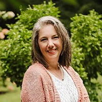 Laura Stone stands in front of green foliage. She is wearing a white top and a peach-colored cardigan. Facing the camera and smiling, the owner of Bearded Lady Soap Factory radiates with her timeless elegance. - Market Design Team: Define. Structure. Expand.