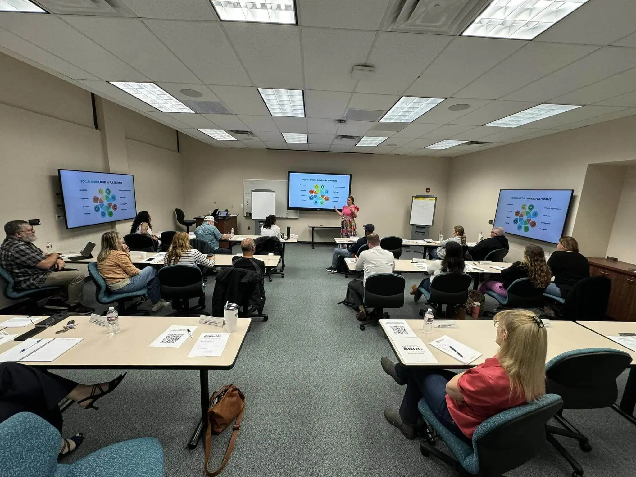 A classroom at Brazosport College Small Business Development Center. Margarey Valdez of MDT is presenting near a screen displaying "Social Media Digital Platforms." Several other screens around the room show the same diagram. Papers and notebooks are on the tables. - Market Design Team: Define. Structure. Expand.