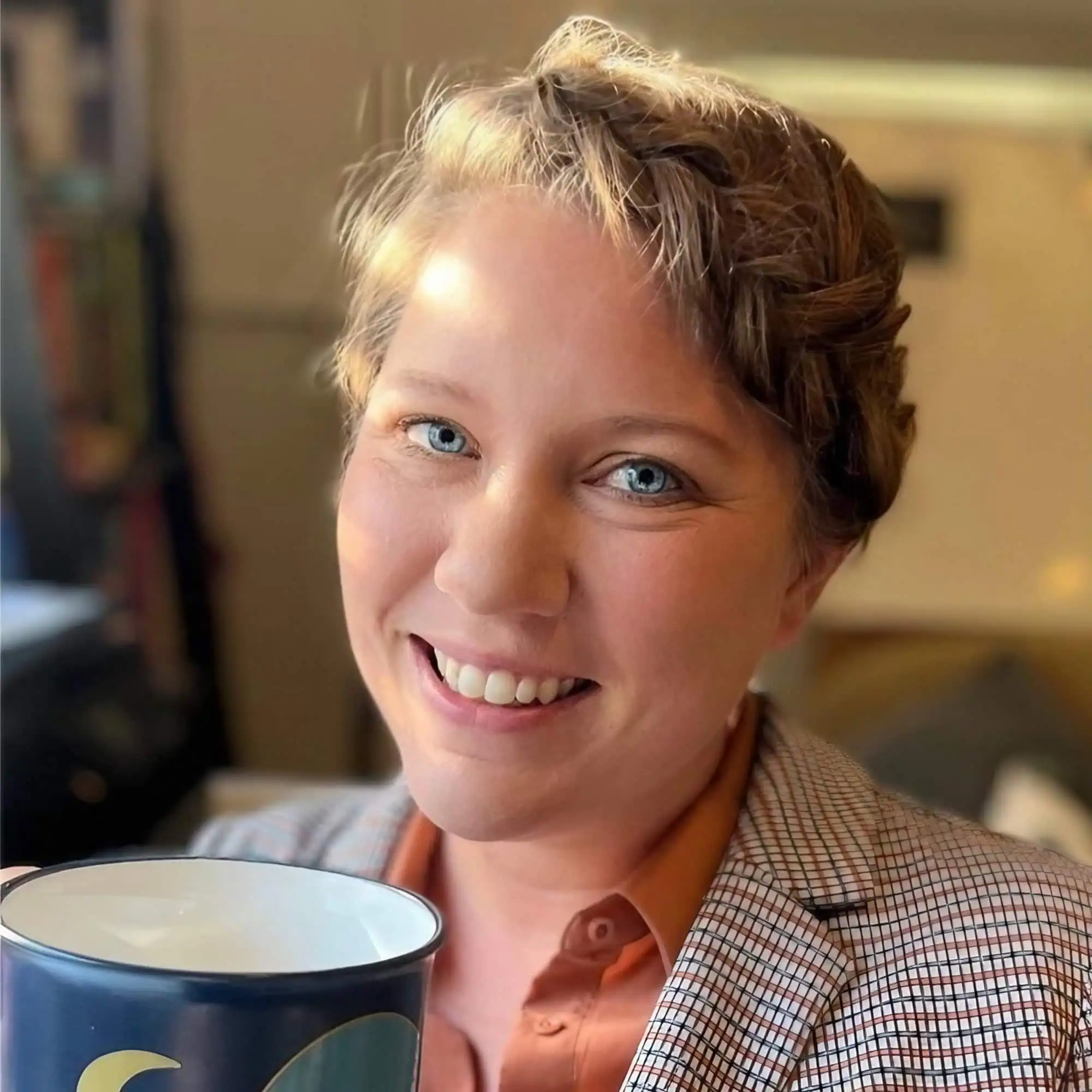 A person with short curly hair is smiling at the camera while holding a coffee mug. They are wearing a plaid blazer and an orange shirt, reminiscent of Margarey Valdez's signature style. The background appears to be an indoor setting with warm lighting. - Market Design Team: Define. Structure. Expand.
