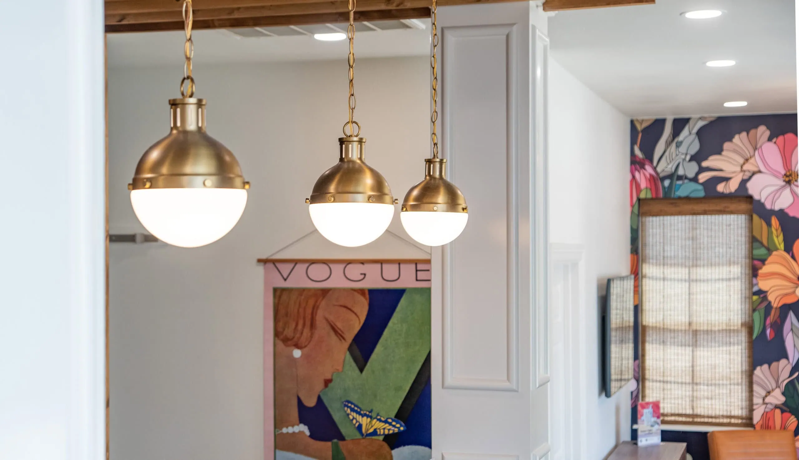 Three spherical pendant lights with brass finishes hang from the ceiling, creating a well-curated look. A colorful art piece featuring a profile of a woman and the word "Vogue" complements the urban vibe of the room, while floral-patterned wallpaper adorns part of the space. - Market Design Team: Define. Structure. Expand.