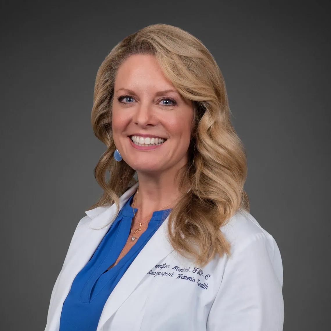 A woman with long blonde hair is smiling, wearing a blue blouse beneath her pristine white lab coat. Her blue earrings and necklace subtly catch the light. The lab coat, embroidered with indecipherable text, stands out against the plain dark gray backdrop, reflecting an urban style of elegance. - Market Design Team: Define. Structure. Expand.