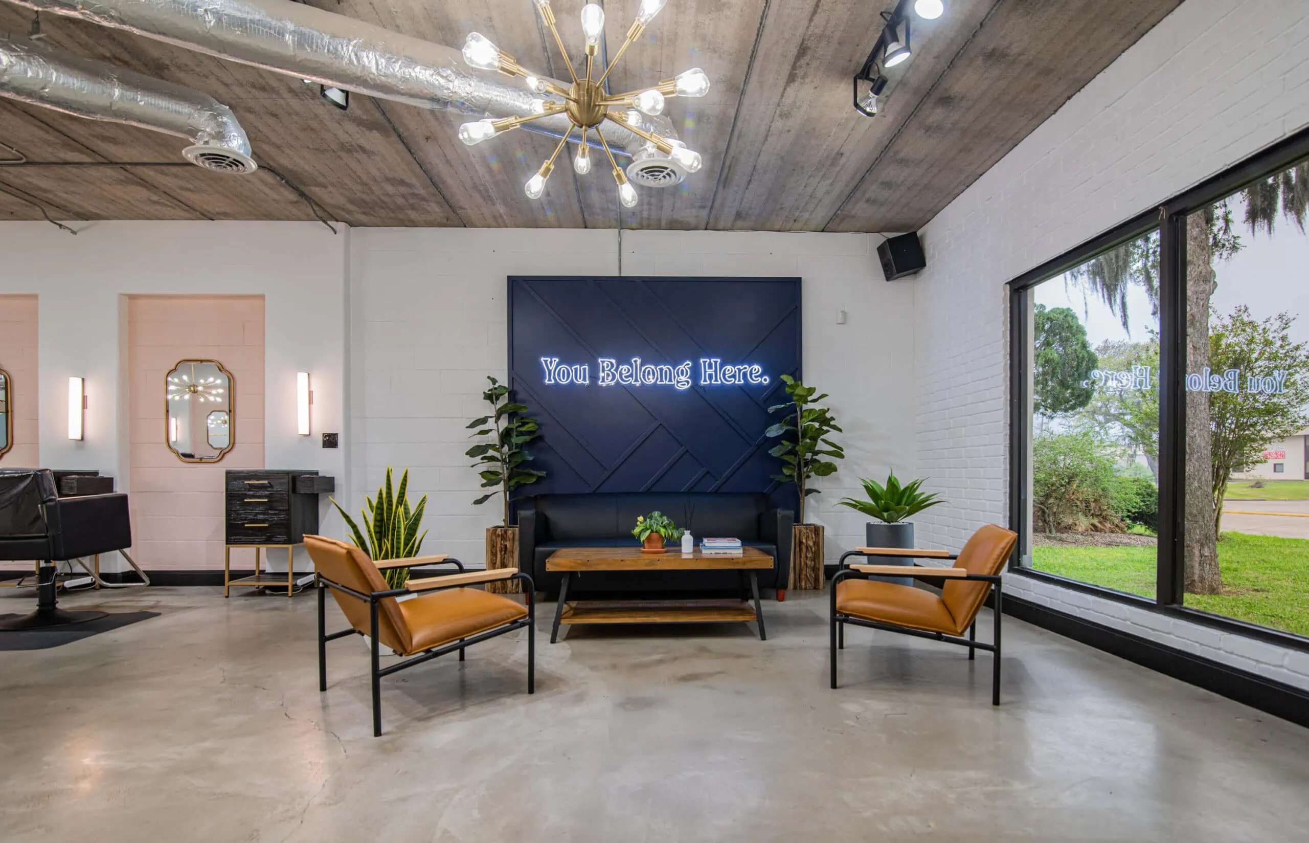 A modern waiting area at Urban Eve Salon features a black sofa, two tan leather chairs, and a wooden coffee table. A neon sign reads "You Belong Here" above the sofa. The boutique space has potted plants, exposed ceiling ducts, and a large window showcasing greenery outside. - Market Design Team: Define. Structure. Expand.