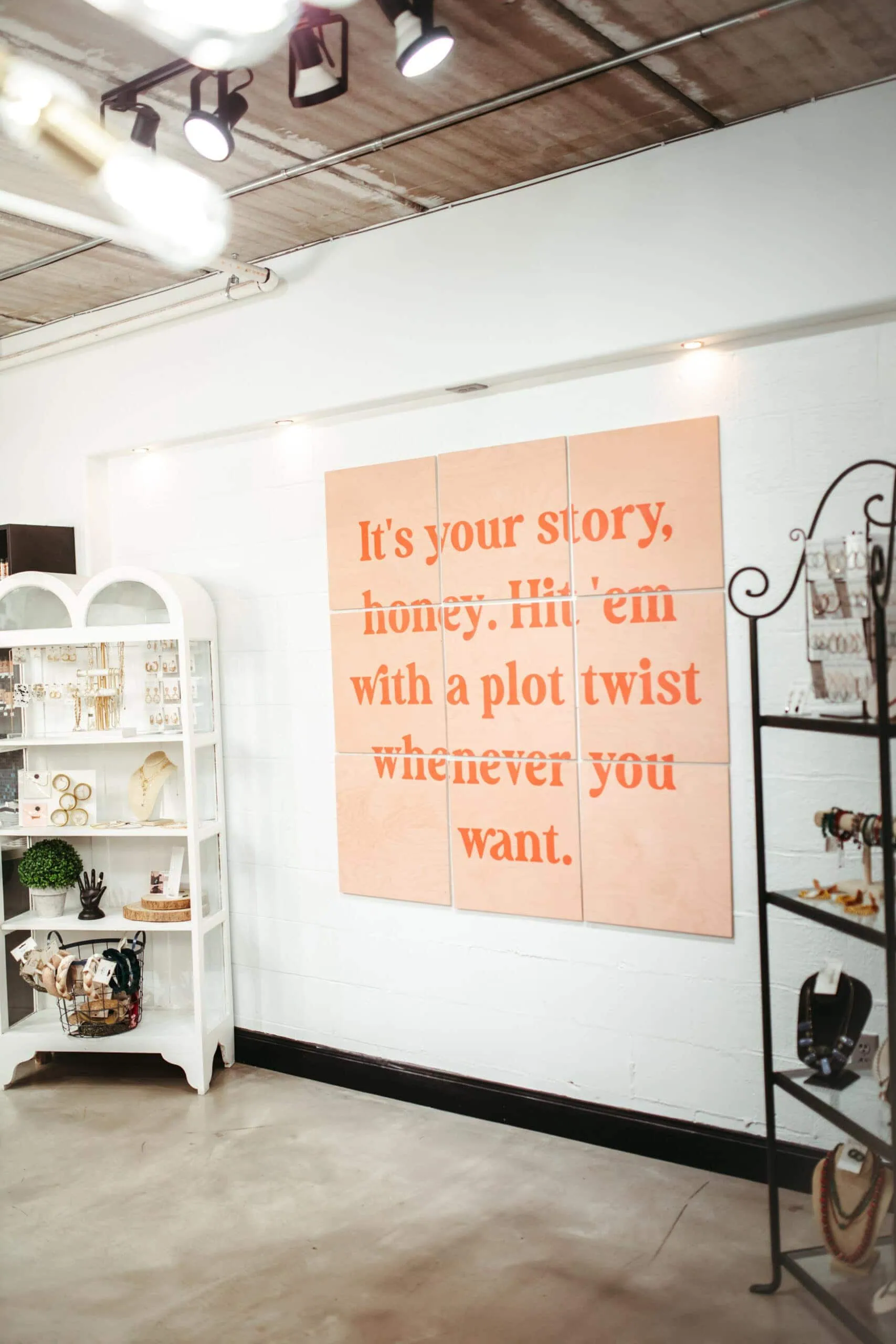 A well-lit room features a white bookshelf on the left displaying decor items and a black metal stand on the right showcasing jewelry. The wall sign reads, "It's your story, honey. Hit 'em with a plot twist whenever you want." Perfect for an Urban Eve Salon & Boutique ambiance. - Market Design Team: Define. Structure. Expand.