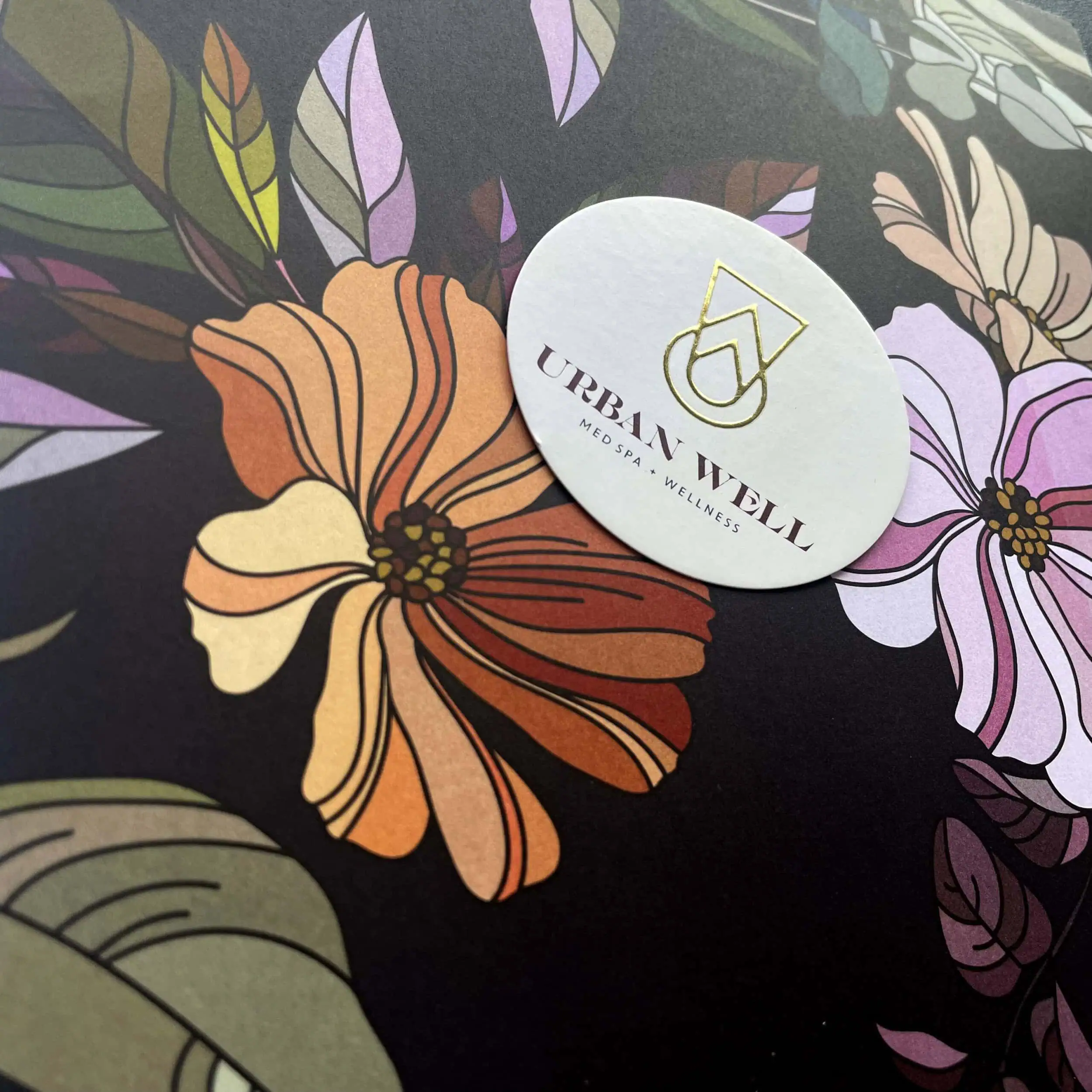 A close-up of a floral-patterned surface featuring stylized orange and pink flowers with green leaves. A white circular sticker with gold text reads "Urban Well Med Spa + Wellness. - Market Design Team: Define. Structure. Expand.