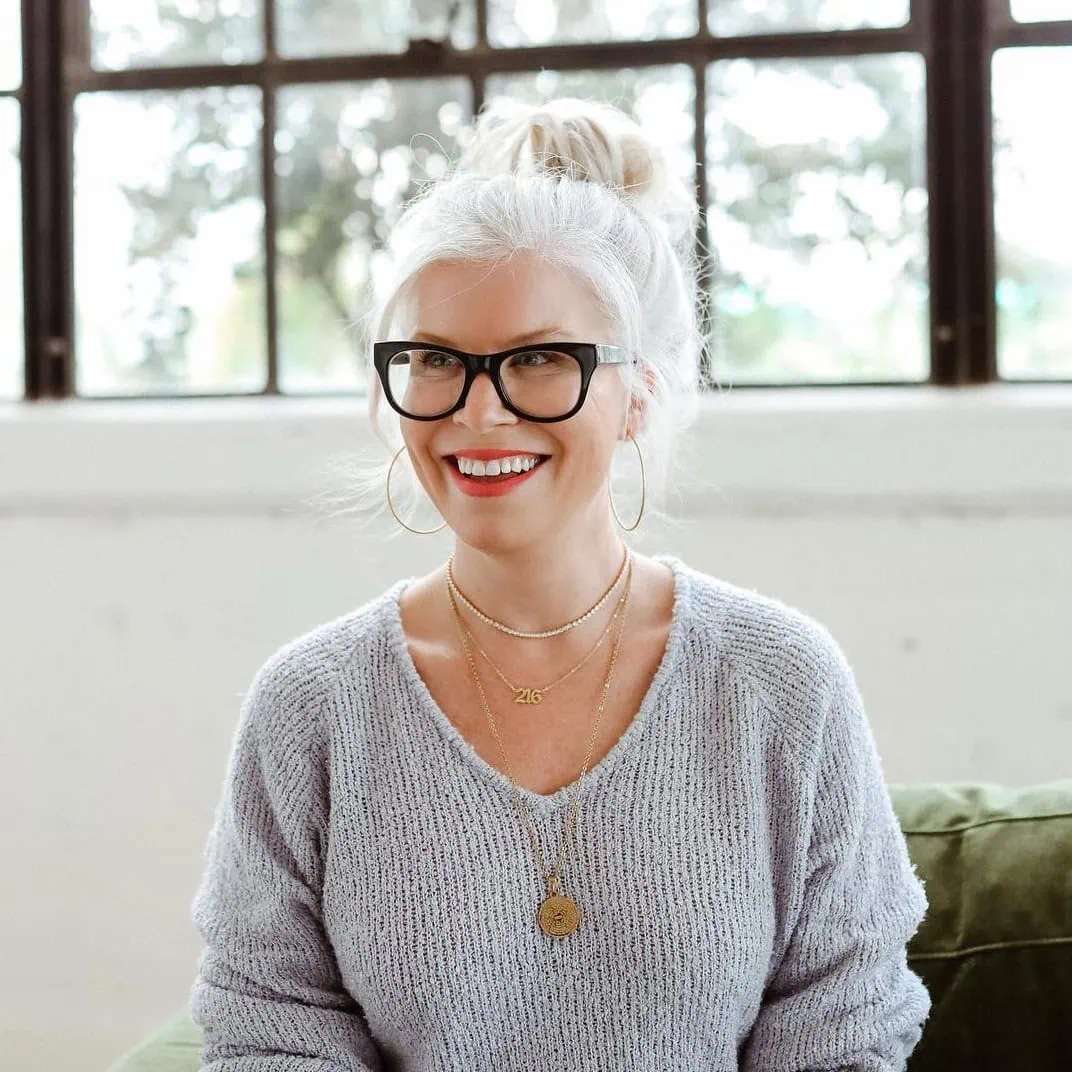 A woman with white hair in a bun, wearing glasses and hoop earrings, smiles while sitting. Her light gray sweater and layered necklaces give her a chic appearance, reminiscent of someone who just stepped out of Urban Eve Salon. The background shows a window with a blurred view of trees outside. - Market Design Team: Define. Structure. Expand.