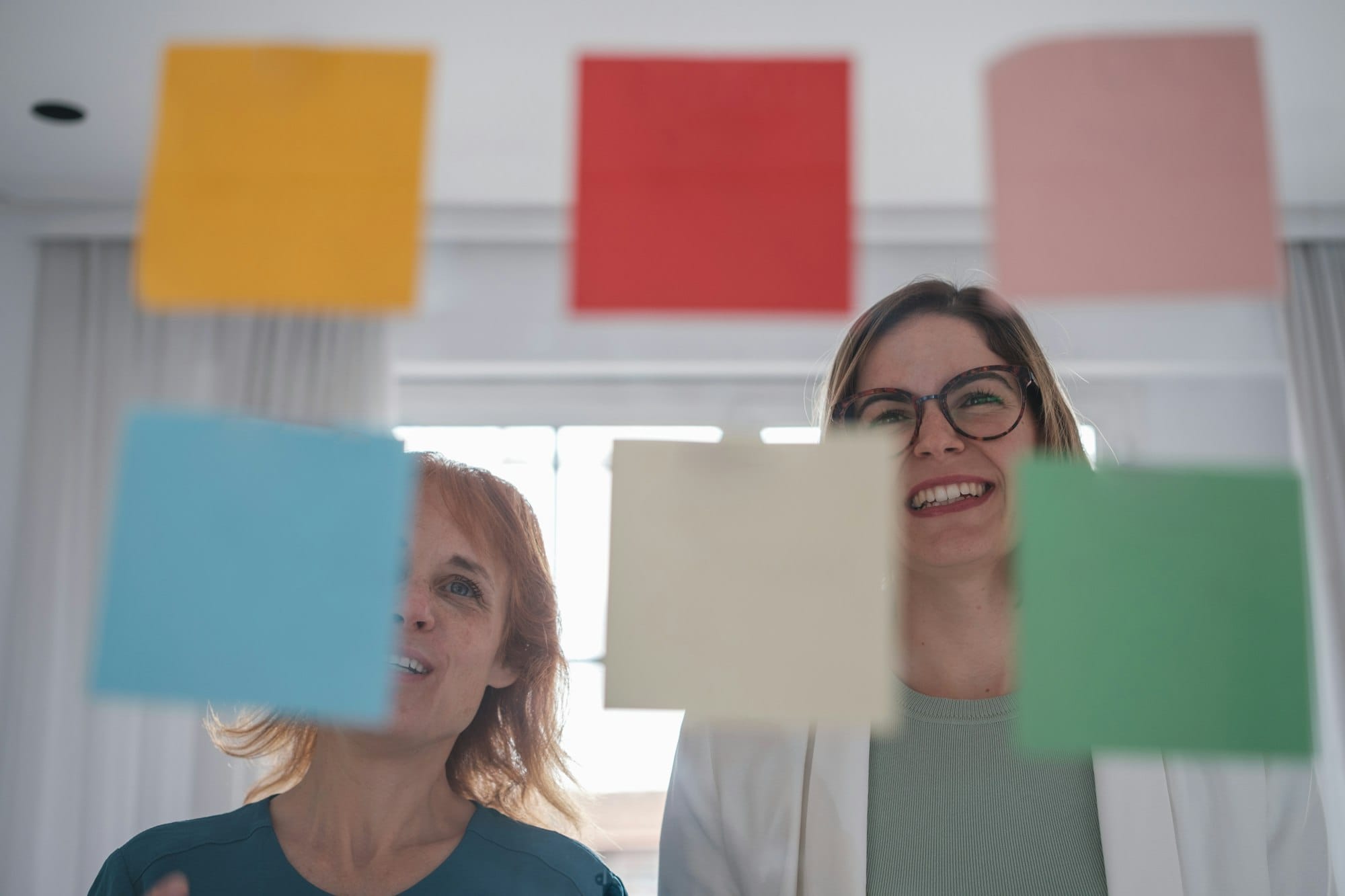 Two women looking at their workplace blackboard with post-it notes and working together. Small business planning.