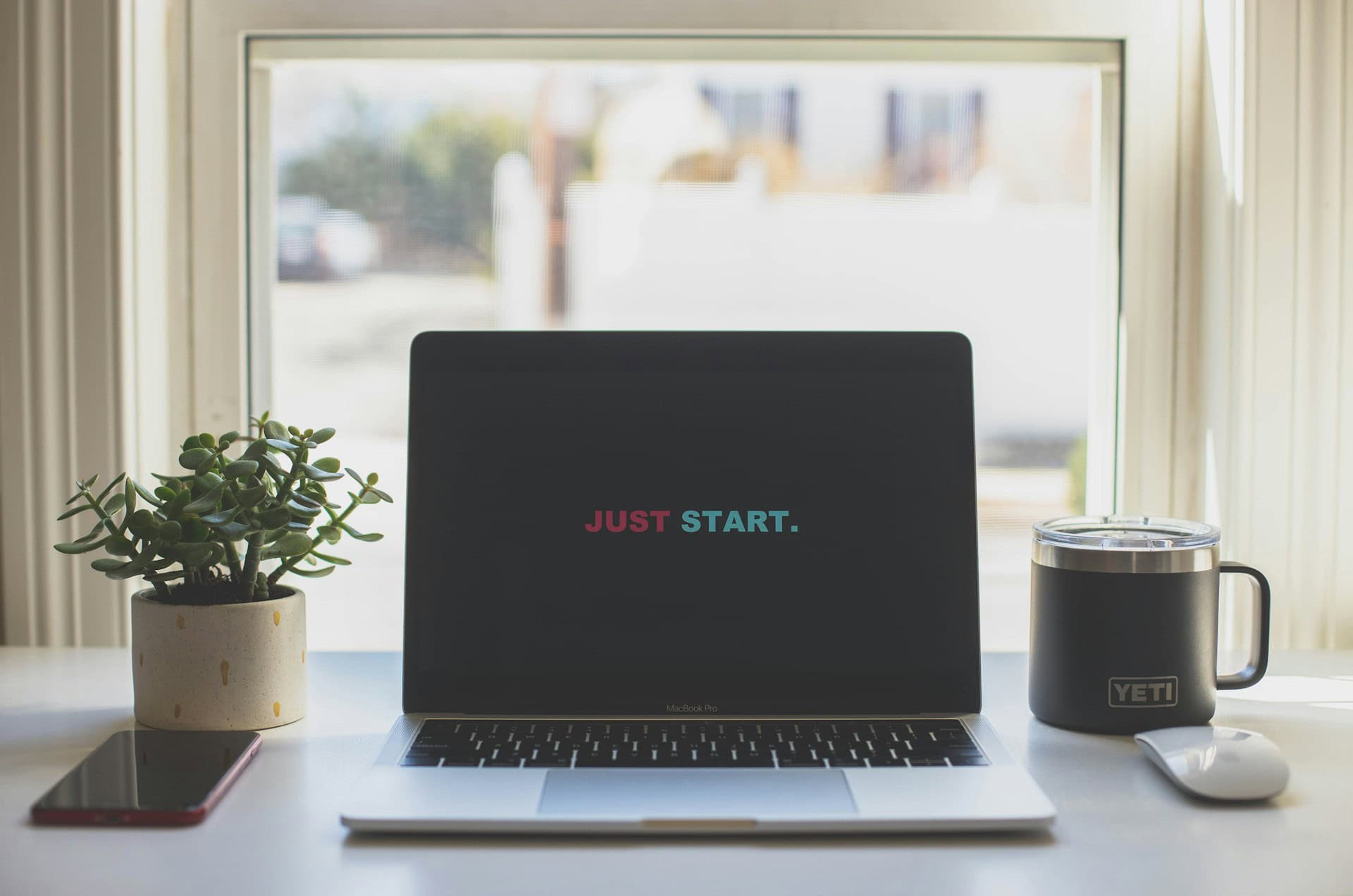 A laptop on a desk displays the words "JUST START" on its screen, hinting at new beginnings in website hosting. Next to it, a small potted plant sits left, while a Yeti-brand mug rests right. A smartphone lies beside the plant with a window as its backdrop. - Market Design Team: Define. Structure. Expand.