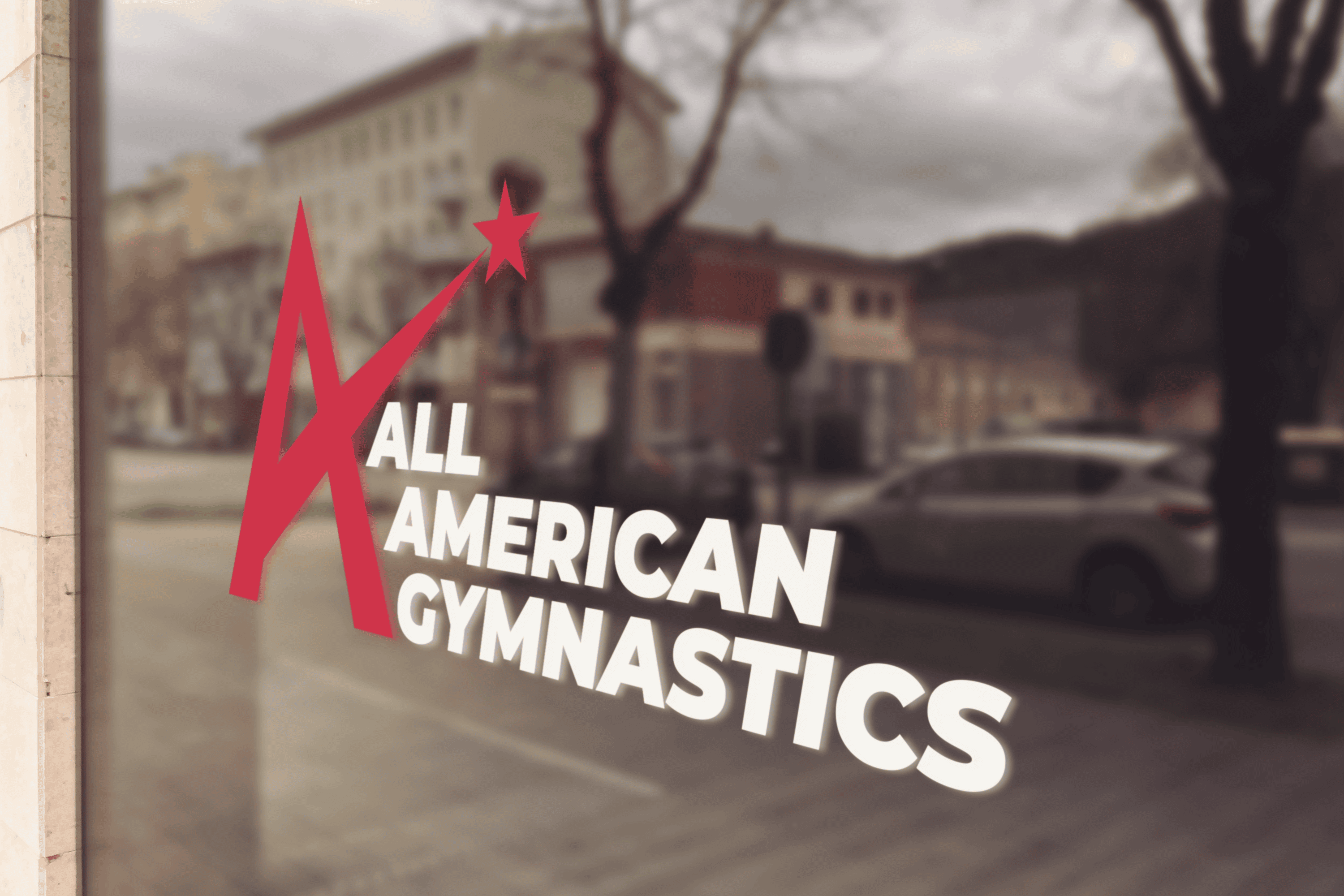 A window sign reads "All American Gymnastics" with a large red letter "A" forming a star. The sign is on glass, reflecting buildings, cars, and trees from the street outside. - Market Design Team: Define. Structure. Expand.