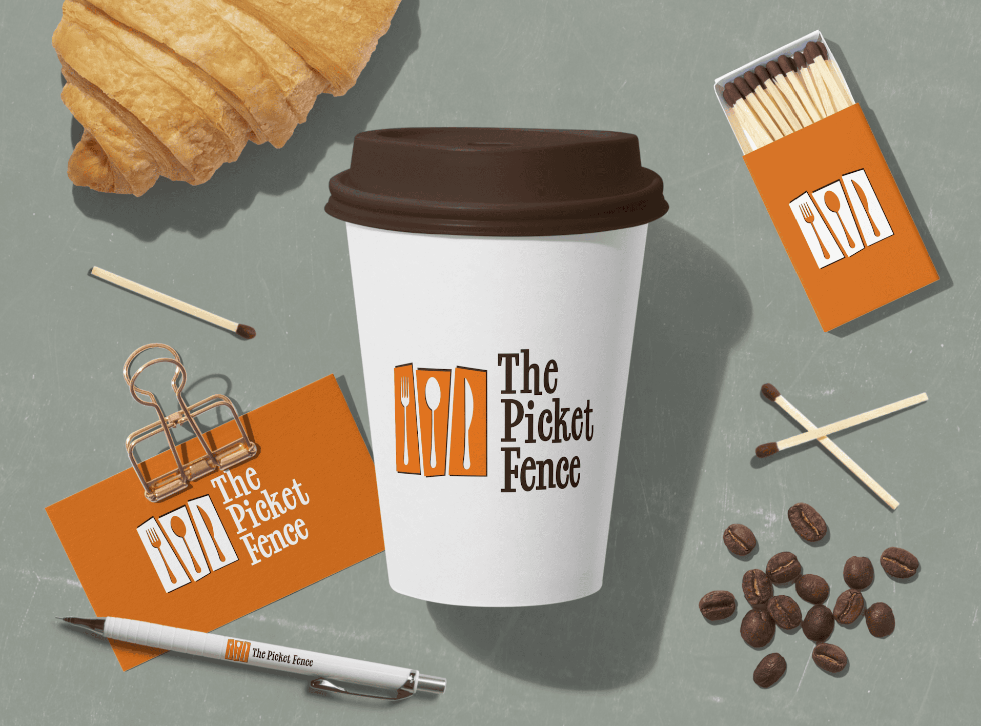 A coffee cup with "The Picket Fence" logo is centered on a gray surface, surrounded by a croissant, matchbox, matches, business card, binder clip, pen, and coffee beans, all branded with the same logo. - Market Design Team: Define. Structure. Expand.