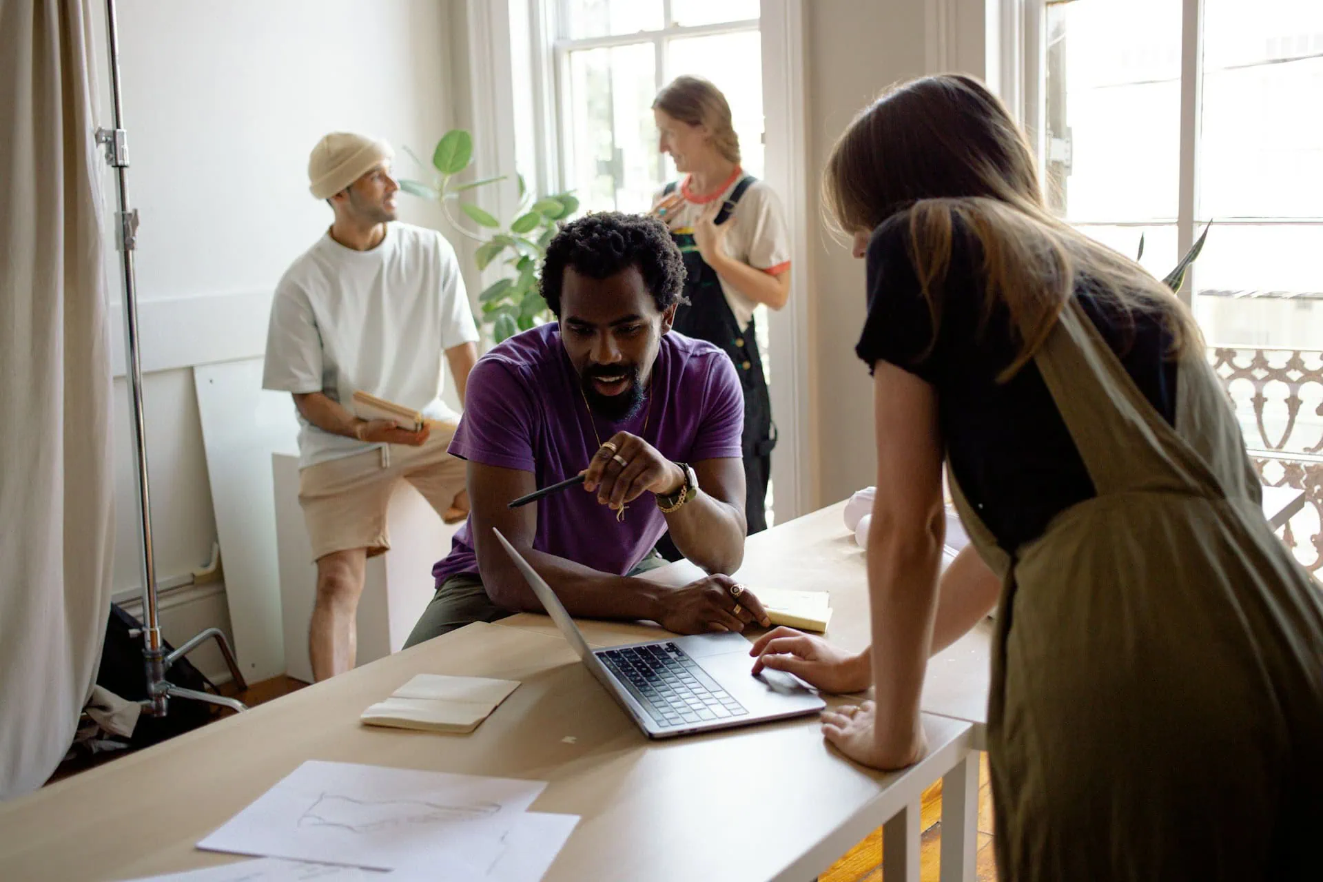 Four people in a bright office space collaborate around a desk, working with laptops and notes. Two discuss ideas on a laptop, while the others talk in the background—just another productive day for leading graphic design companies. - Market Design Team: Define. Structure. Expand.
