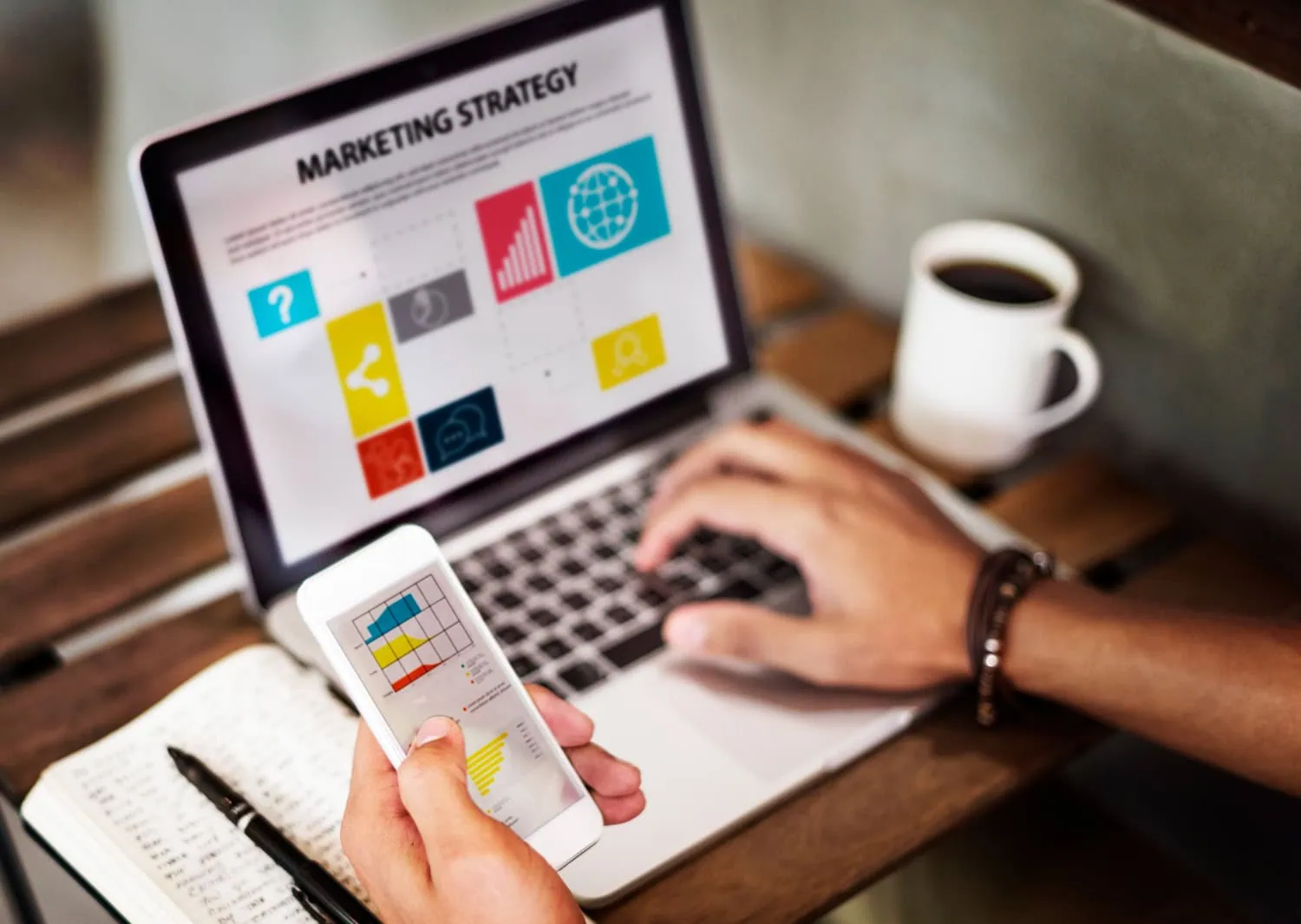 A person works on a laptop displaying a digital marketing strategy presentation with charts and icons, while holding a smartphone showing a graph. A notebook and cup of coffee sit on the wooden table, capturing a productive fall day in Lake Jackson. - Market Design Team: Define. Structure. Expand.
