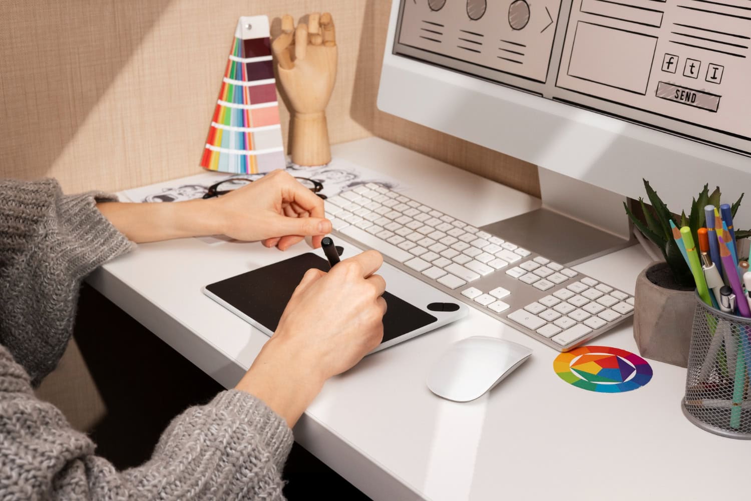 A person works on a digital drawing tablet at a white desk in Lake Jackson, focused on graphic design. A computer displays a design mockup, with color swatches, stationery, a wooden mannequin hand, and a color wheel coaster nearby. - Market Design Team: Define. Structure. Expand.