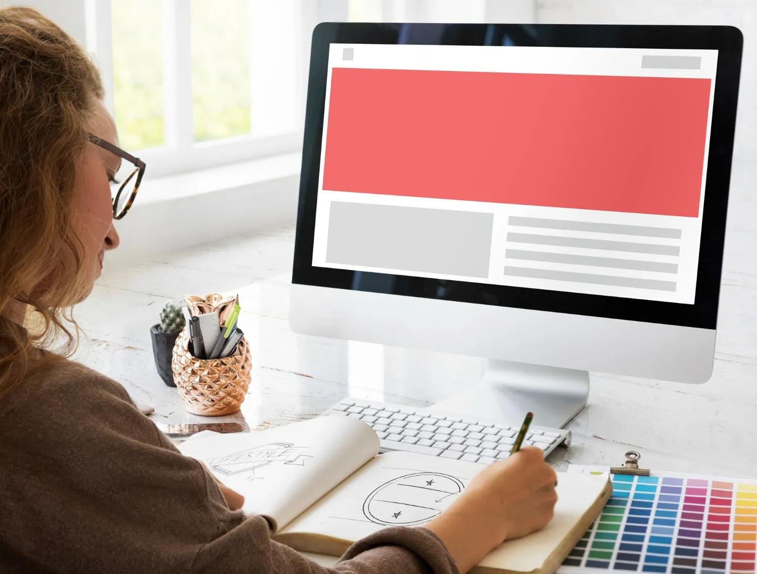 A person sits at a desk sketching in a notebook, with a color palette and computer monitor displaying a rural web design layout. Natural light streams through a nearby window, highlighting the creative process. - Market Design Team: Define. Structure. Expand.