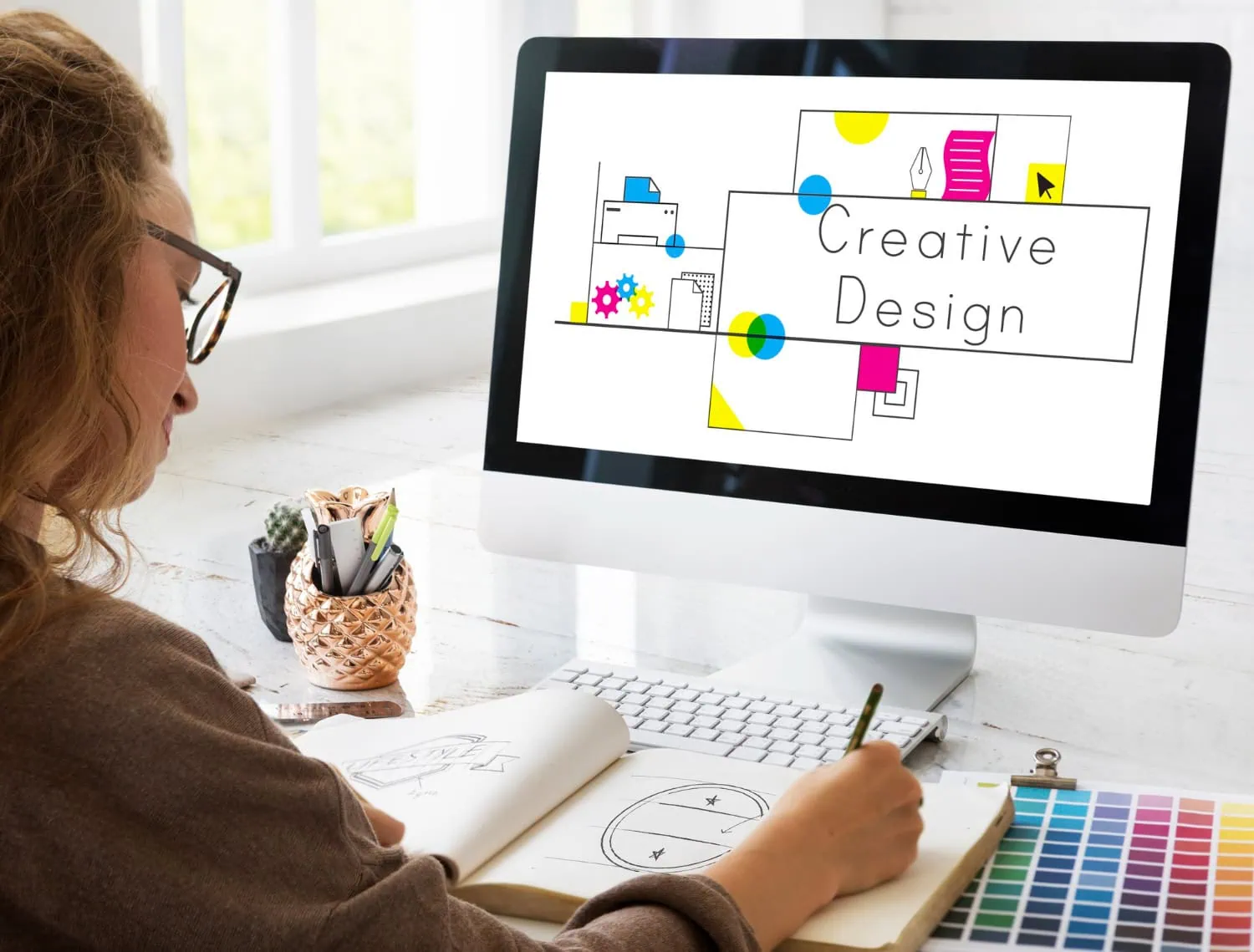 A person with long hair and glasses sketches in a notebook at a desk. A computer monitor displays a colorful design with the text "Creative Design." Art supplies and a plant are on the desk. - Market Design Team: Define. Structure. Expand.