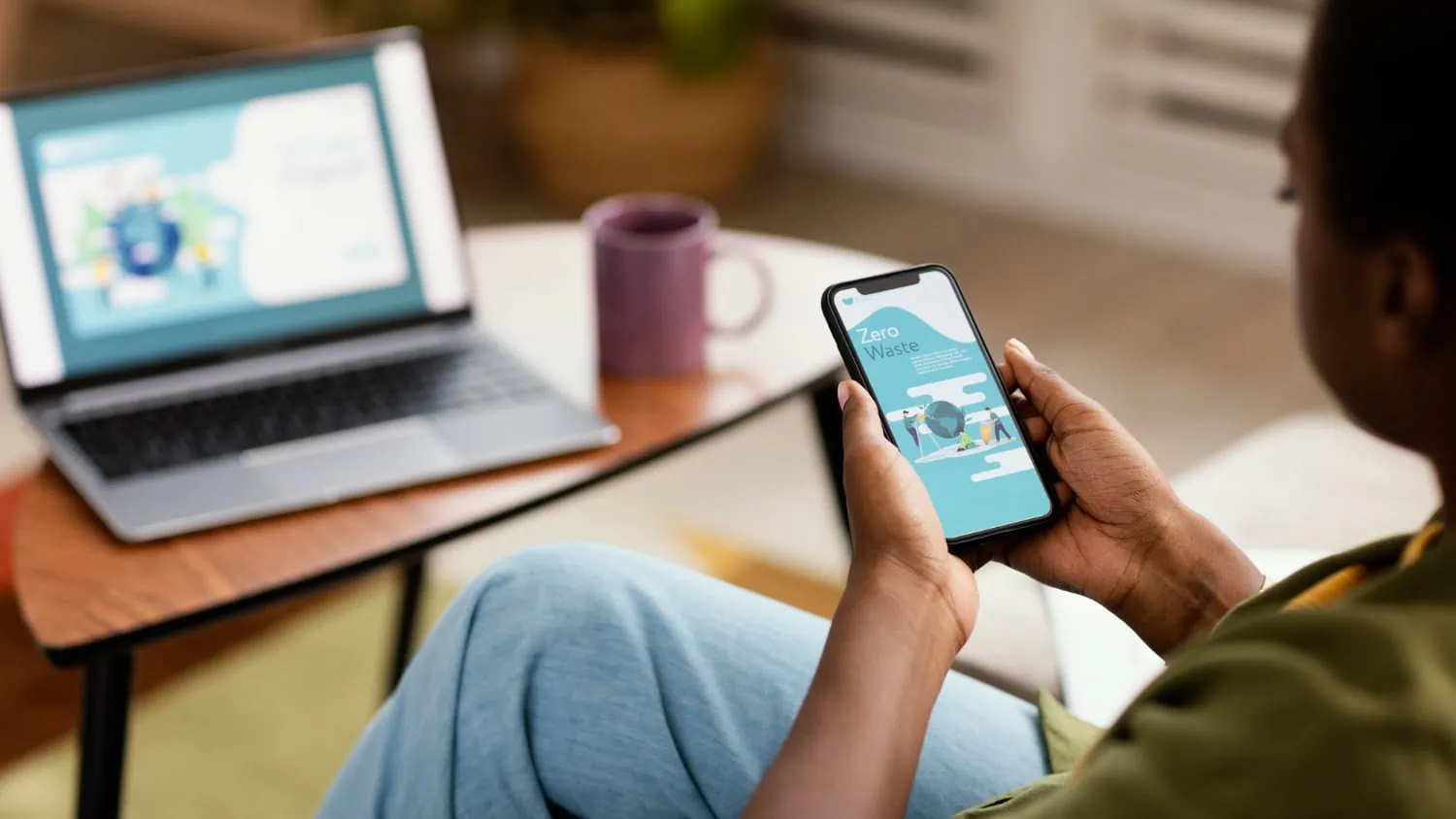 A person sits holding a smartphone displaying a "Zero Waste" screen, with a laptop nearby showing website tips on the same topic and a purple mug next to it. The cozy indoor setting highlights the mobile first website approach. - Market Design Team: Define. Structure. Expand.