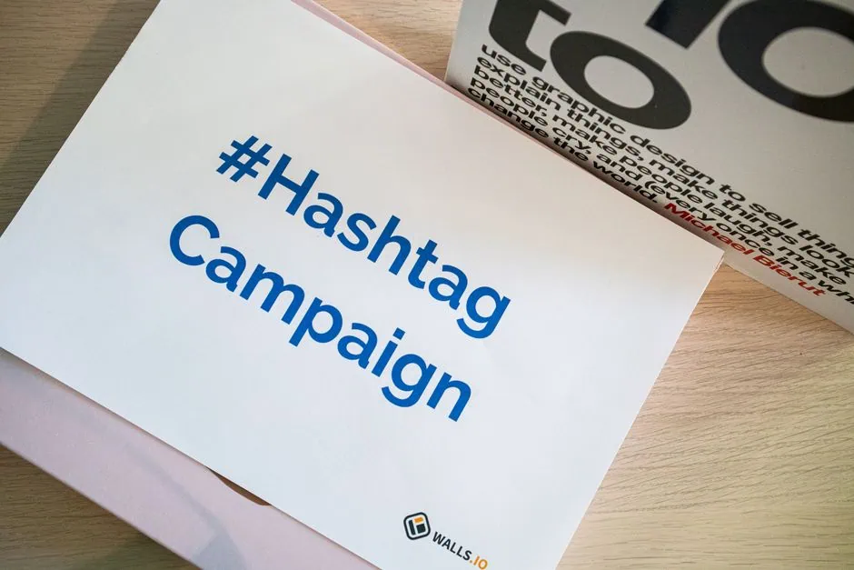 a sheet of paper on a wooden surface displays the text “#hashtag campaign” in blue letters, with a small walls.io logo in the bottom center. the clean layout showcases thoughtful graphic design, making it ideal for social media or use as part of a practical guide. part of another printed item appears in the background. market design team: define. structure. expand.