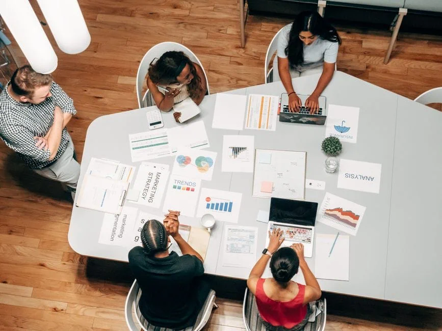 five people are seated around a table with laptops, charts, and printed materials, engaged in a collaborative work session focused on business seo strategies. the image is taken from above, capturing a modern office setting with wooden floors. market design team: define. structure. expand.