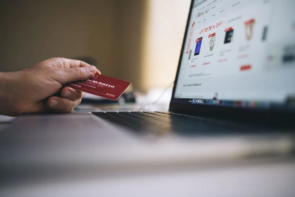a person holds a red credit card in front of a laptop displaying an online shopping website with sales promotions and product images, highlighting the importance of website conversions as emphasized by marketdesignteam.com. market design team: define. structure. expand.