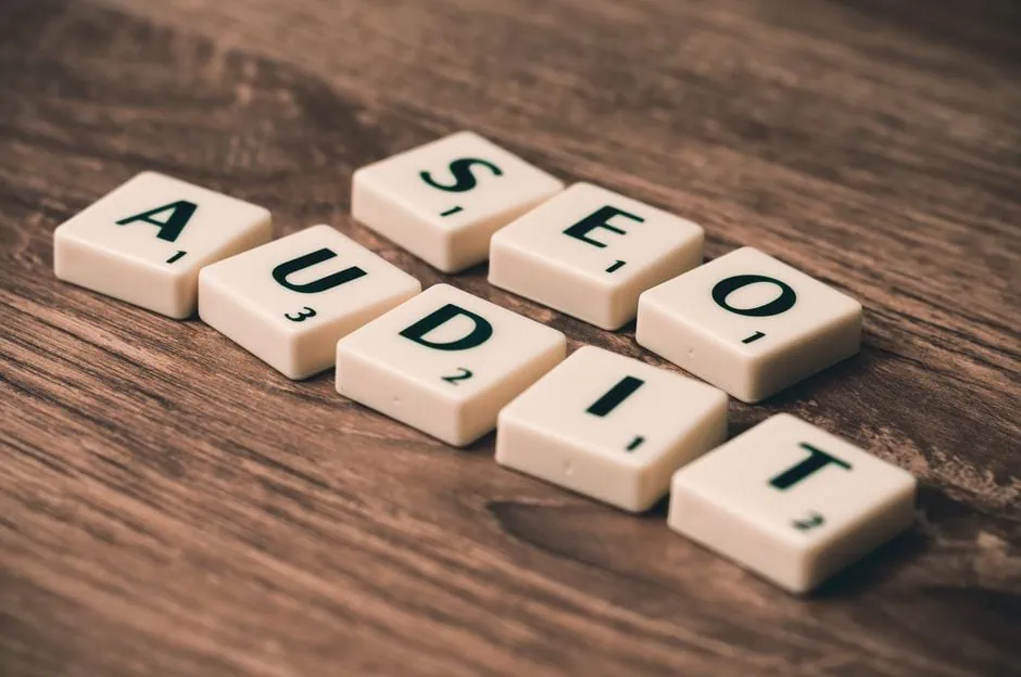 white scrabble tiles on a wooden surface spell out “seo audit,” highlighting seo services essential for any business. the tiles display letter values in the bottom right corners. market design team: define. structure. expand.