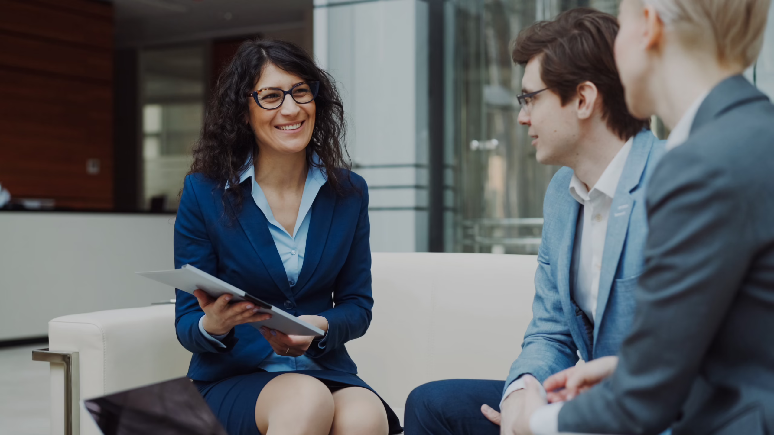 a smiling woman in a navy suit holds a folder while talking with colleagues about hiring a marketing consultant in an office lobby. market design team: define. structure. expand.