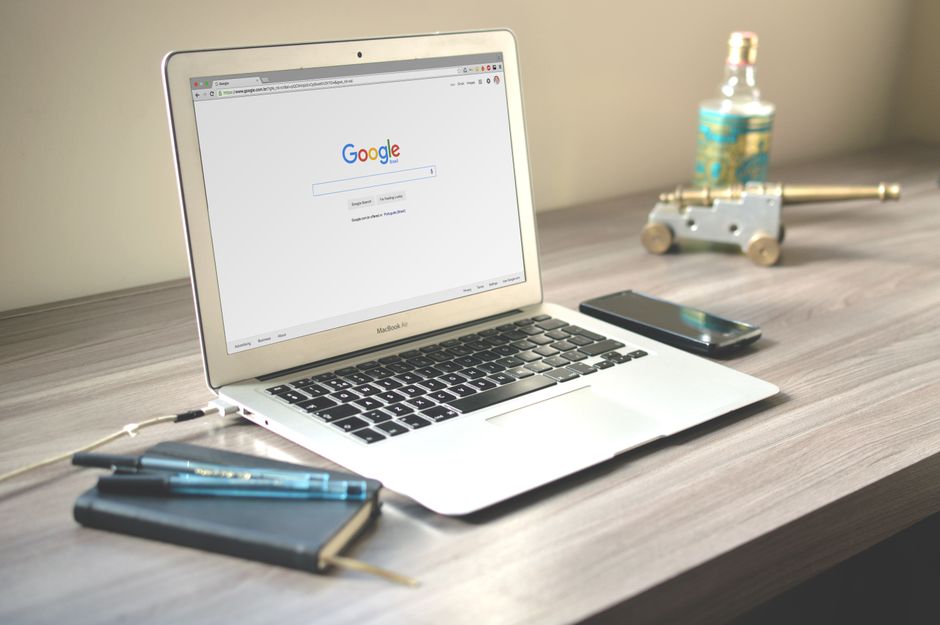 a macbook displaying the google homepage sits on a wooden desk with a notebook, two pens, and a smartphone—perfect for reading a practical guide on seo for small business and getting found online. market design team: define. structure. expand.