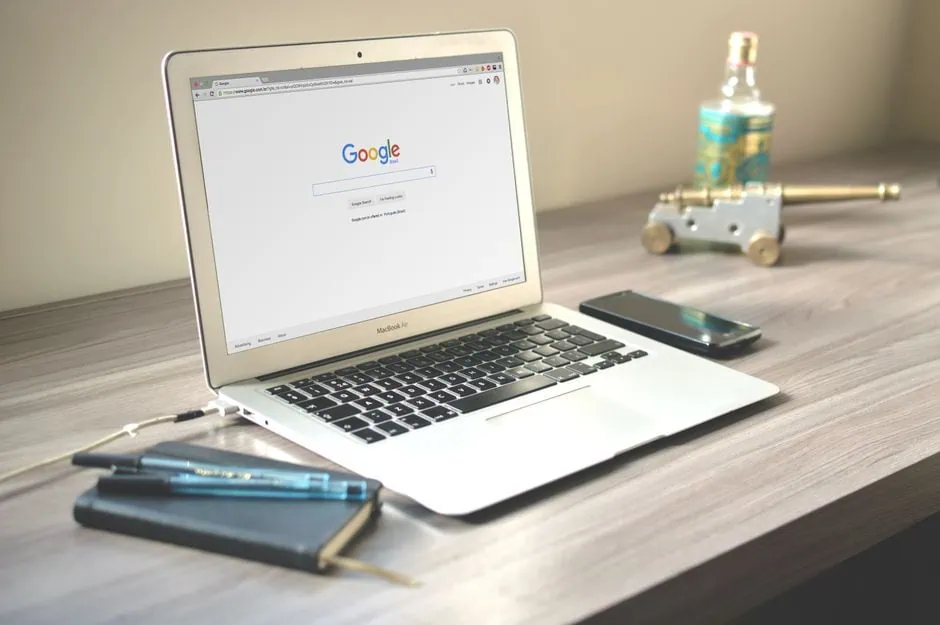 a macbook displaying the google homepage sits on a wooden desk with a notebook, two pens, and a smartphone—perfect for reading a practical guide on seo for small business and getting found online. market design team: define. structure. expand.