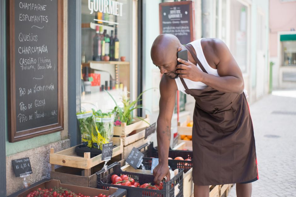a man wearing an apron and tank top talks on the phone while selecting tomatoes from a crate outside his small business produce store. other fruits, vegetables, and a chalkboard menu are visible. market design team: define. structure. expand.
