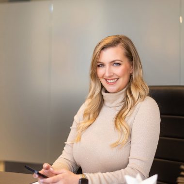 A woman with long blonde hair sits at a desk, smiling at the camera. She is holding a smartphone and wearing a light-colored turtleneck sweater, representing Winnow Financial's approachable and modern style. A closed binder rests on the desk in the professional office setting behind her. - Market Design Team: Define. Structure. Expand.