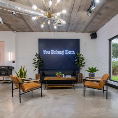 A modern waiting area at Urban Eve Salon features a black sofa, two tan leather chairs, and a wooden coffee table. A neon sign reads "You Belong Here" above the sofa. The boutique space has potted plants, exposed ceiling ducts, and a large window showcasing greenery outside. - Market Design Team: Define. Structure. Expand.